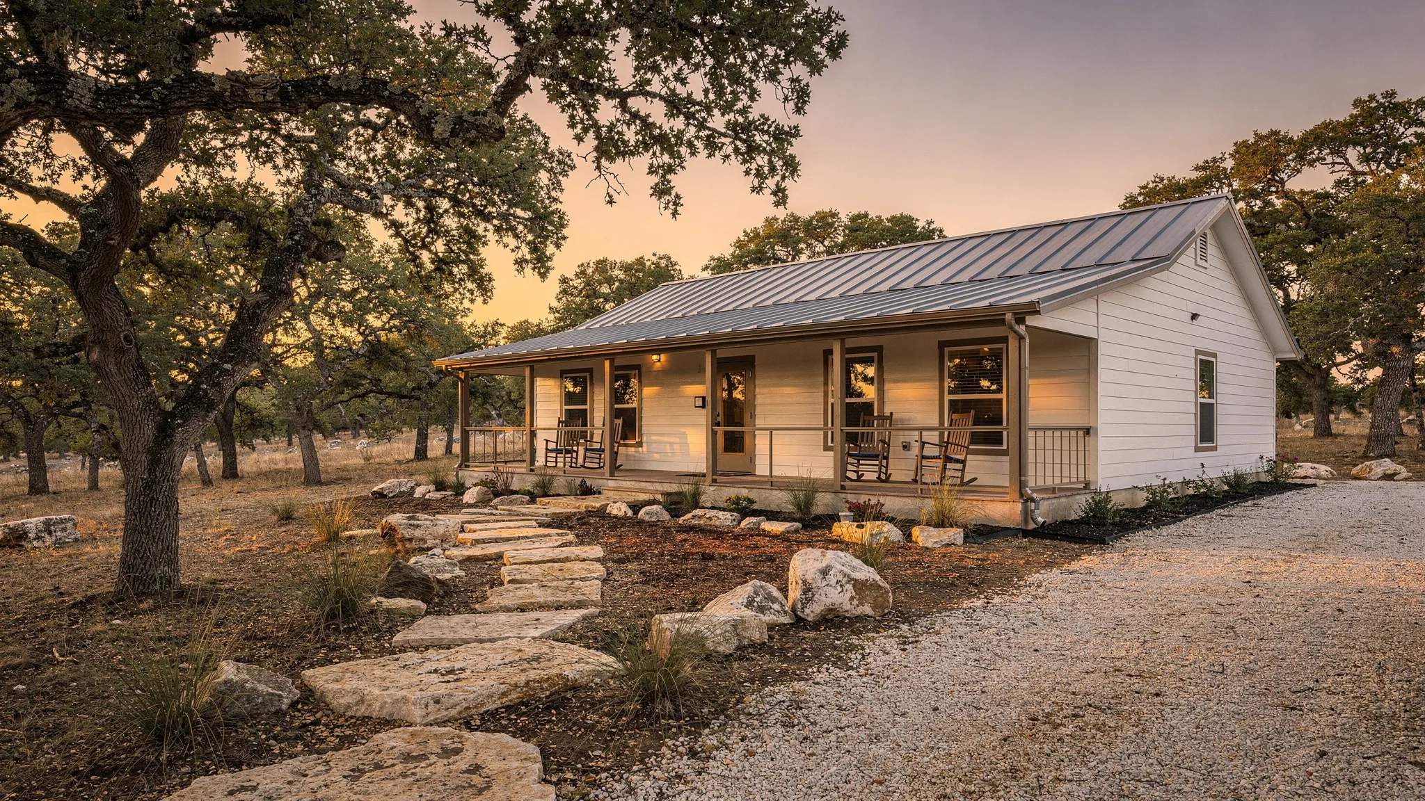 A modern, energy-efficient manufactured home with a wide front porch and metal roof, set on a spacious Texas Hill Country lot near San Antonio with live oaks, limestone steps, and warm evening light.