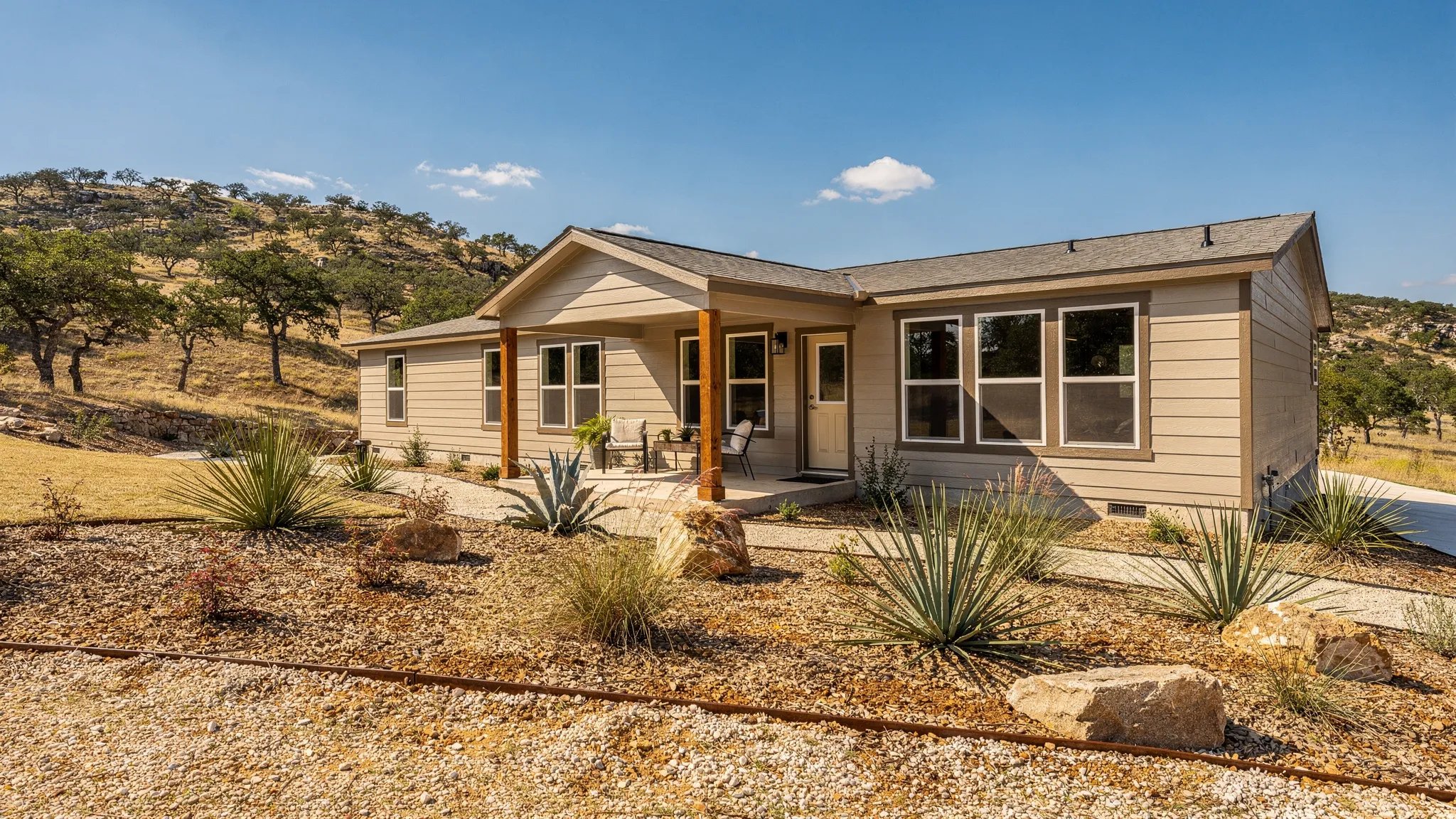 A modern multi-section manufactured home with a covered front porch, xeriscape landscaping, and views of oak-dotted Texas Hill Country under a blue sky.