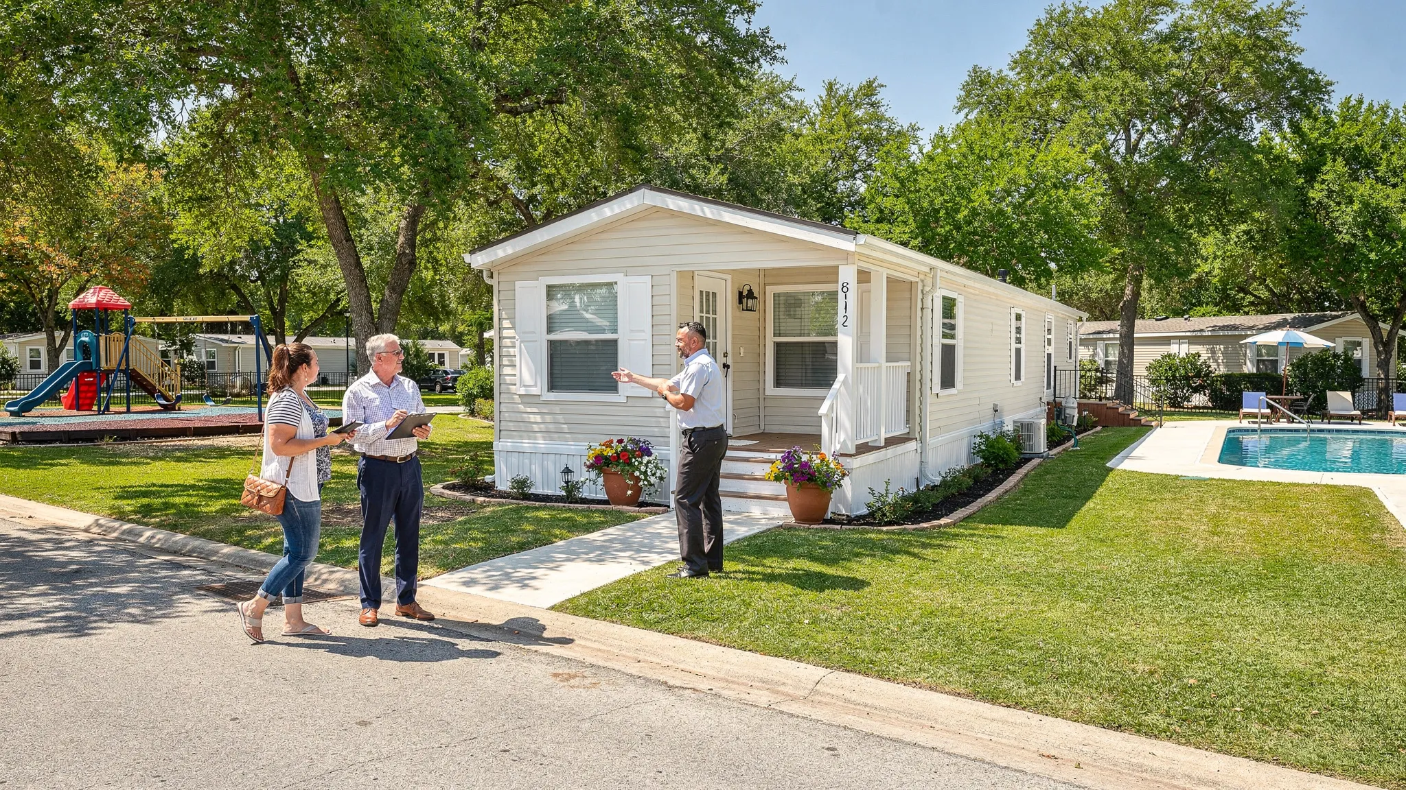A sunny, tree lined manufactured home community street in San Antonio with a modern single wide home, neat skirting, a small front porch with planters, a community pool and playground visible in the background, and a couple touring with a clipboard while a friendly manager greets them.