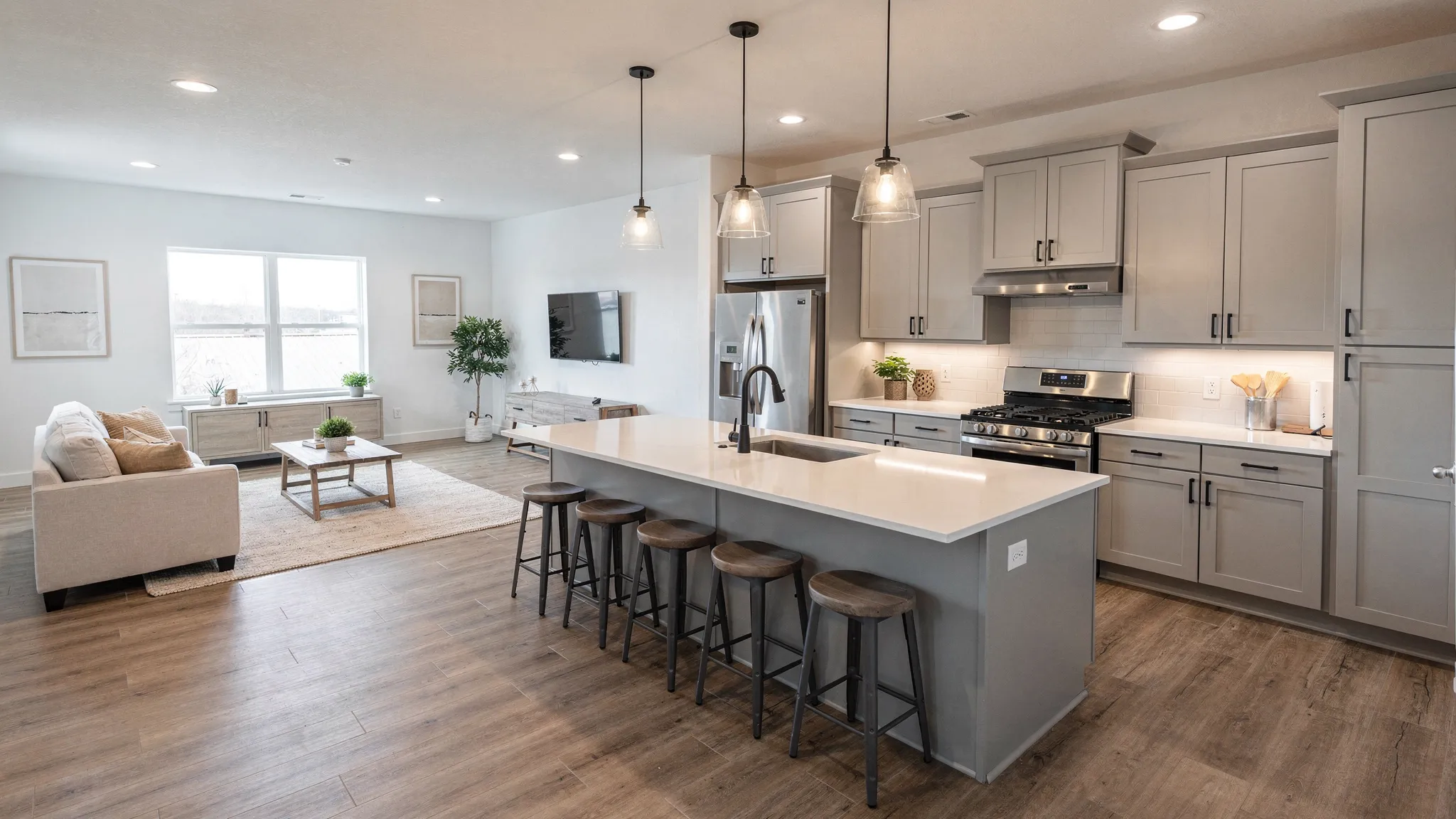 A bright, modern manufactured home interior with an open-concept living room and kitchen, featuring a large kitchen island, shaker-style cabinets, pendant lighting, wood-look flooring, and neutral decor.