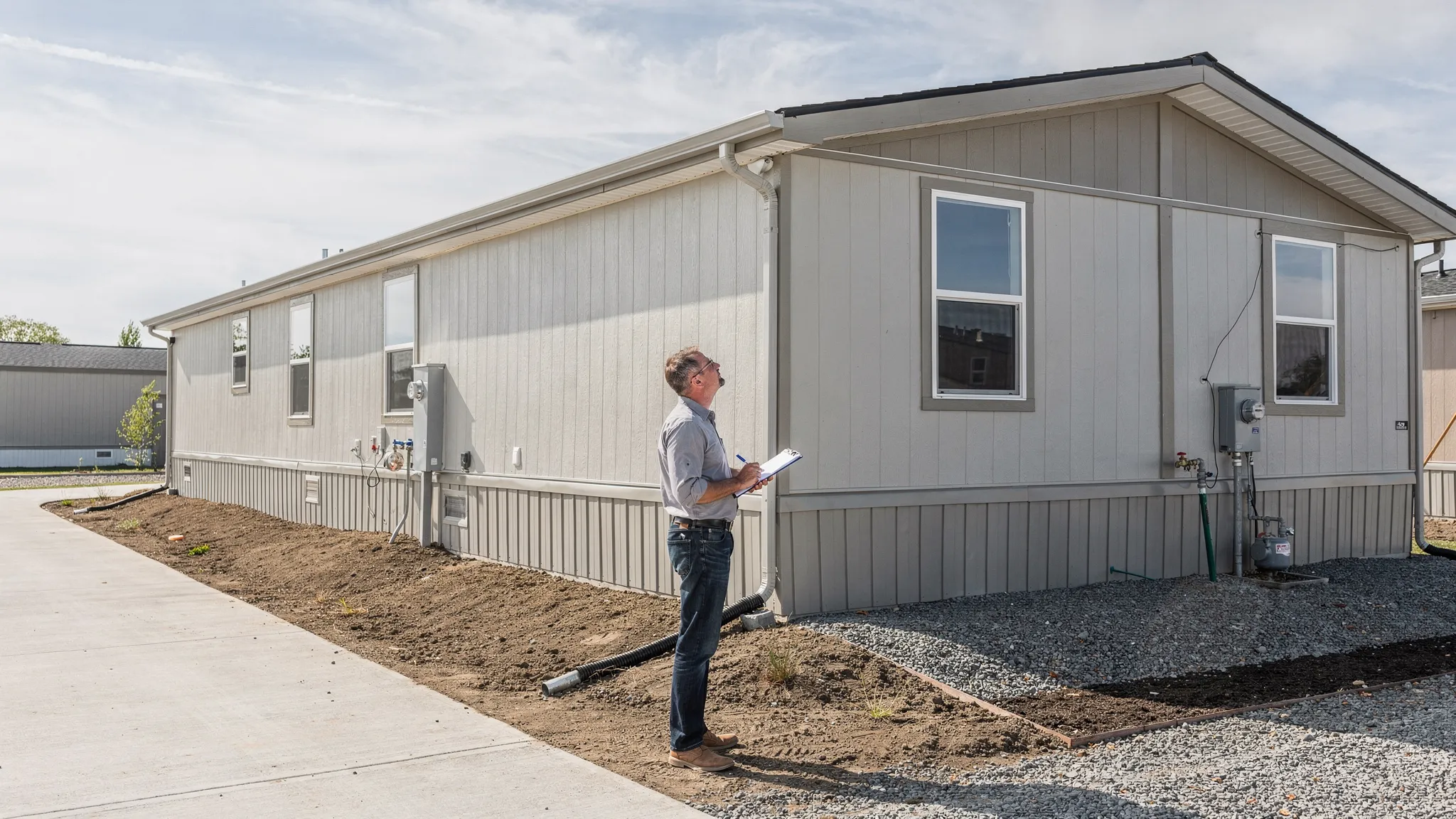 A buyer standing outside a manufactured home holding a clipboard, visually checking the roofline, skirting, and grading around the home, with a clear driveway and utility connections visible.