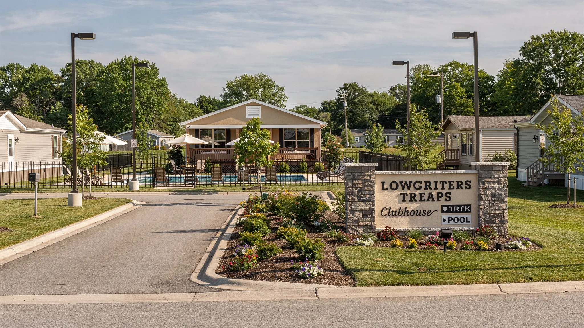 A clean, well-maintained manufactured home community entrance with clear signage, landscaped median, wide internal road, visible street lighting, and a small clubhouse and pool area in the background.
