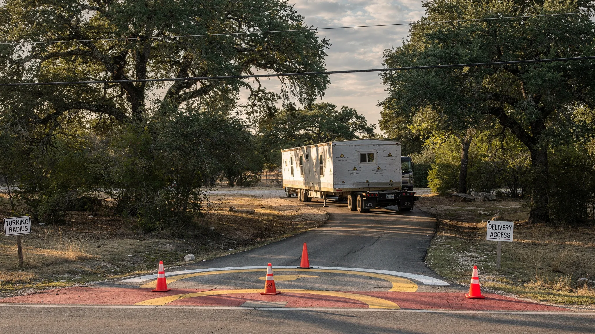 A manufactured home delivery route in a semi-rural San Antonio-area setting, showing a tow vehicle and home section approaching a narrow driveway with trees, overhead utility lines, and a marked turning radius, emphasizing access constraints for installation.