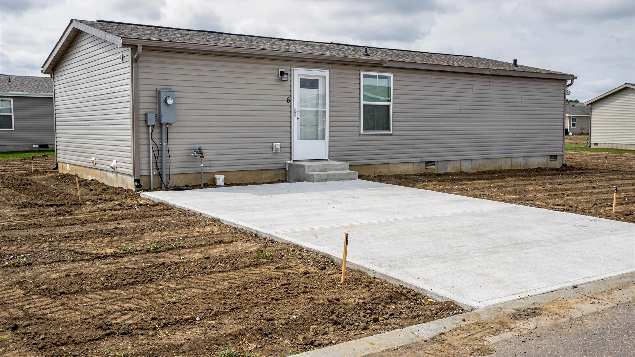 A manufactured home placed on a prepared lot with visible driveway, graded soil, utility hookups (electric meter and water connection), and a clear boundary showing the land parcel.