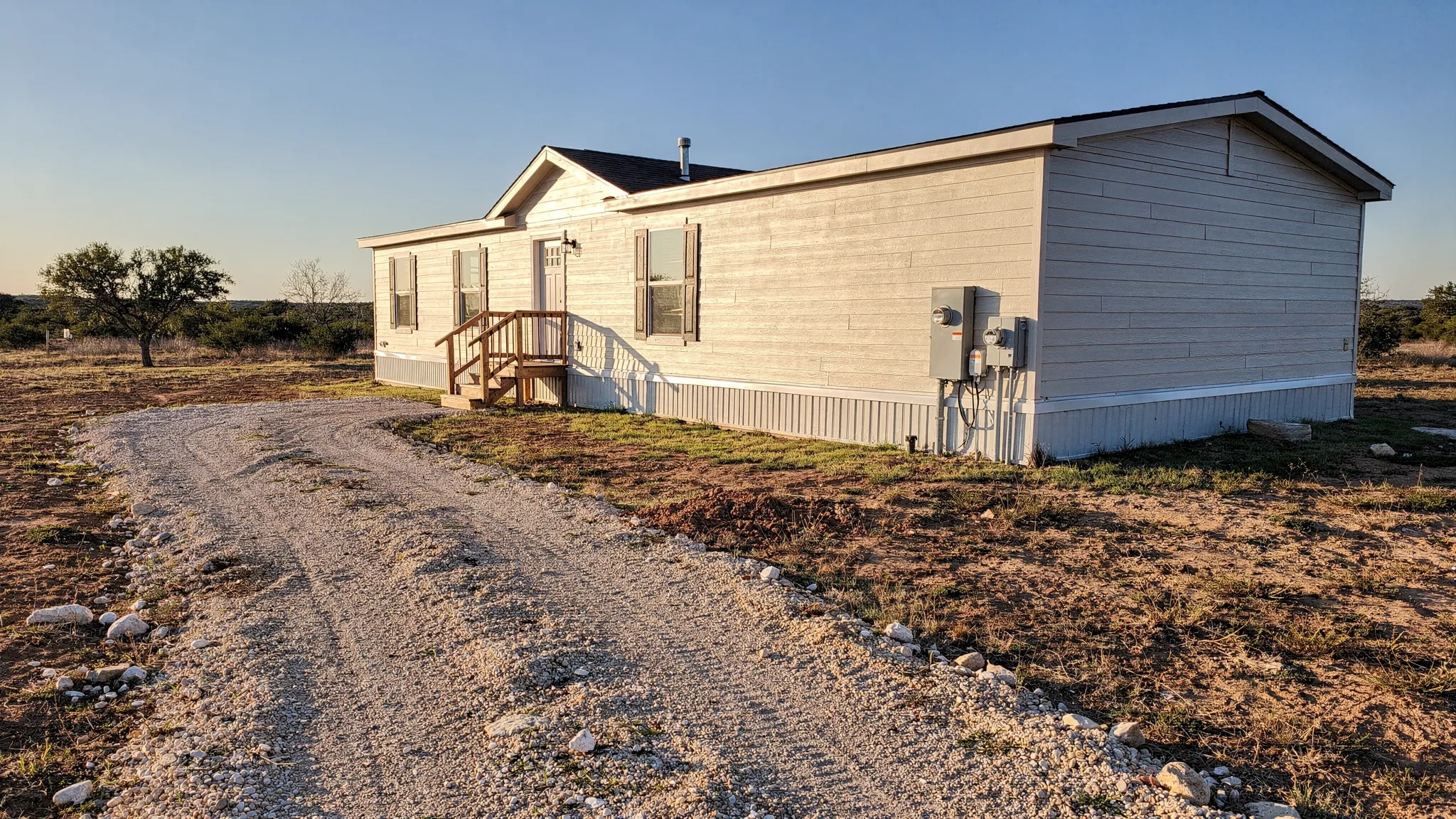 A manufactured home set on a private lot outside San Antonio with a gravel driveway, utility meter, and visible foundation/skirting, showing open space around the home and clear access for delivery.