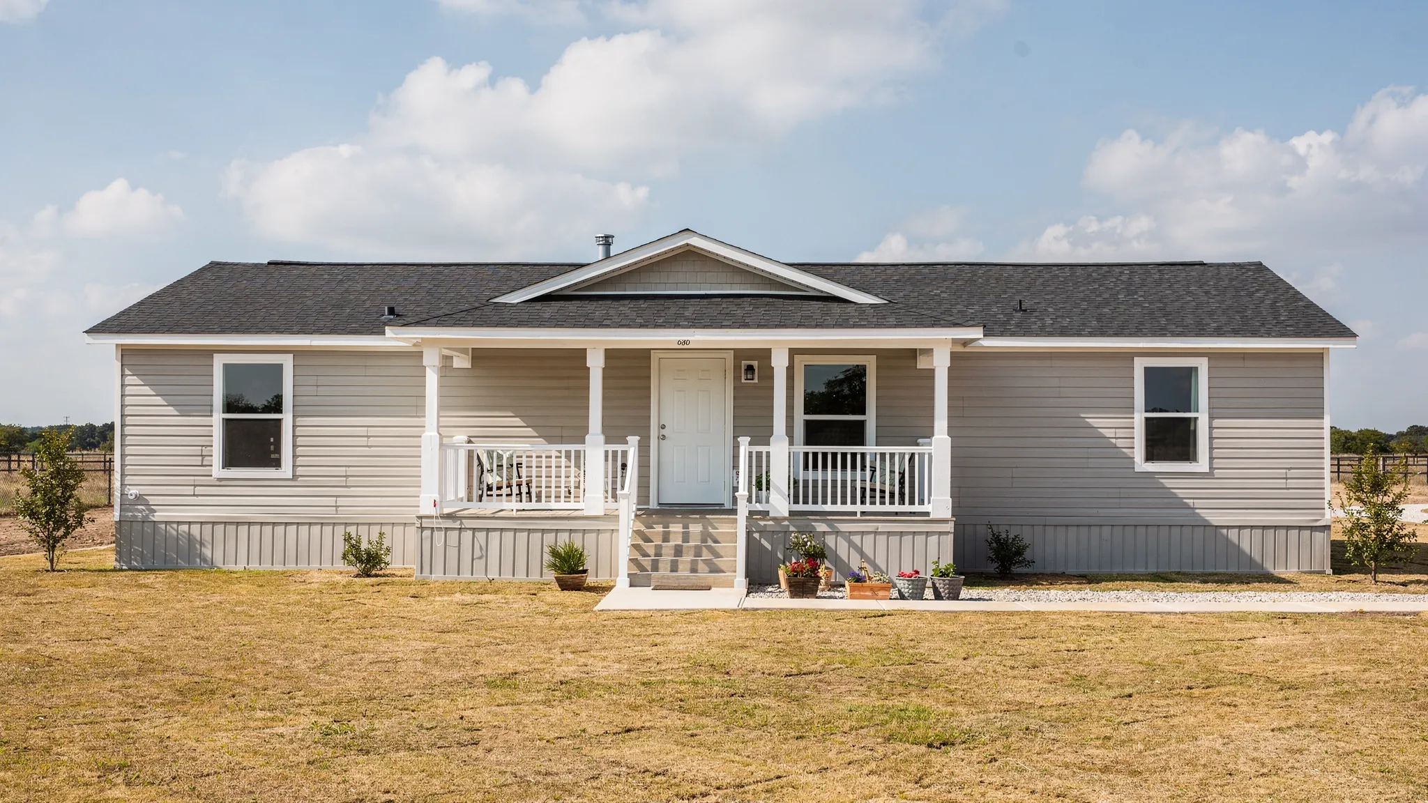 A modern double wide manufactured home exterior in a Texas setting, with a wide front porch, pitched roof, and clean siding lines, shown on a prepared homesite with skirting and steps installed.