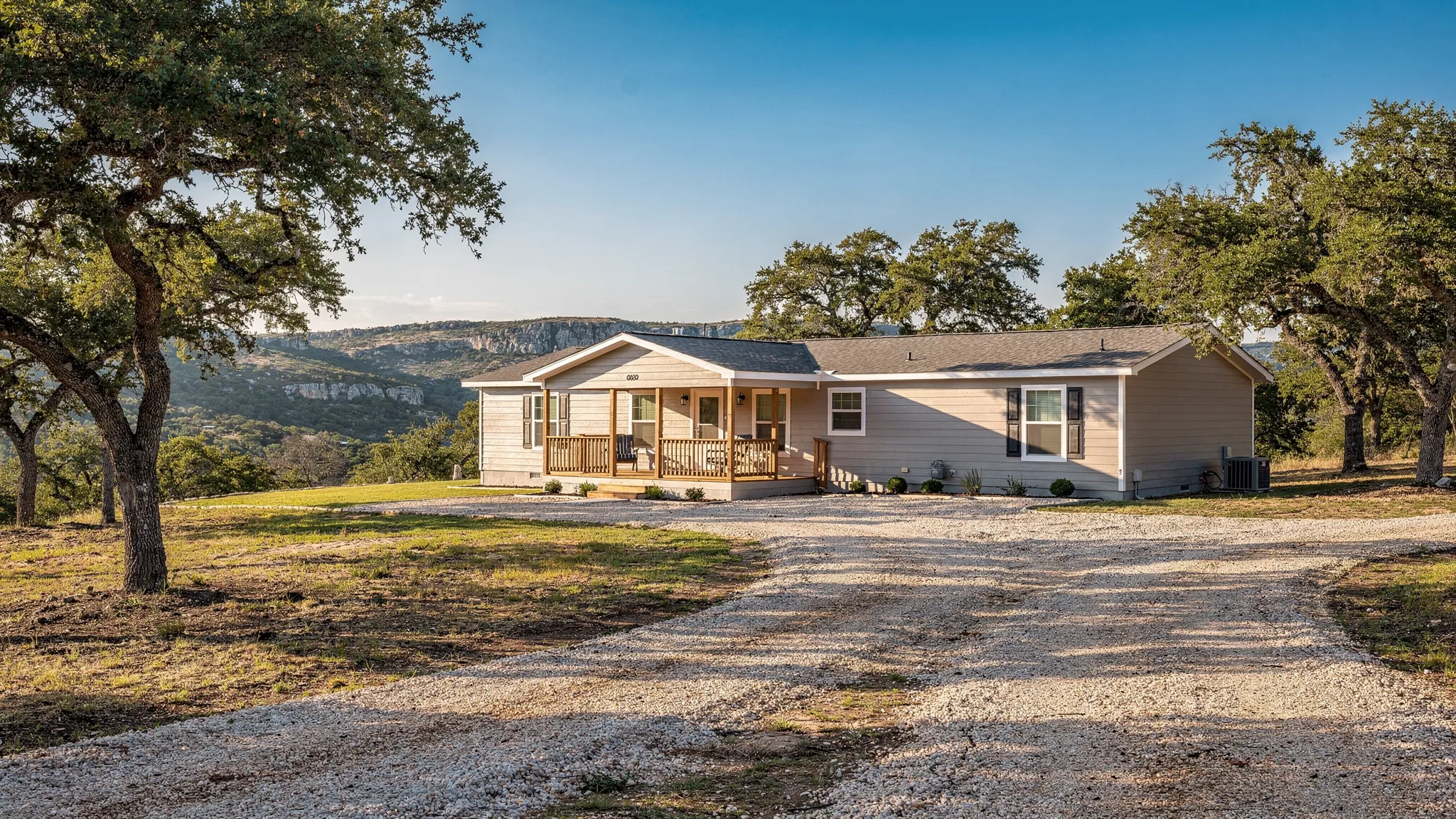 A modern double‑wide manufactured home with a front porch set on 2 acres in the Texas Hill Country near San Antonio, surrounded by live oak trees, crushed granite driveway, and a distant view of rolling limestone hills under a clear summer sky.
