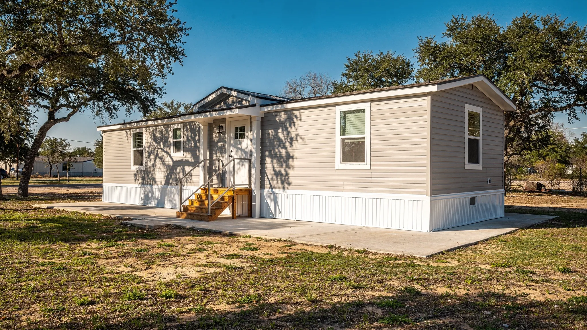 A modern single-section manufactured home installed on a finished foundation with steps and skirting, set on a sunny South Texas lot with mature trees and a clear sky.