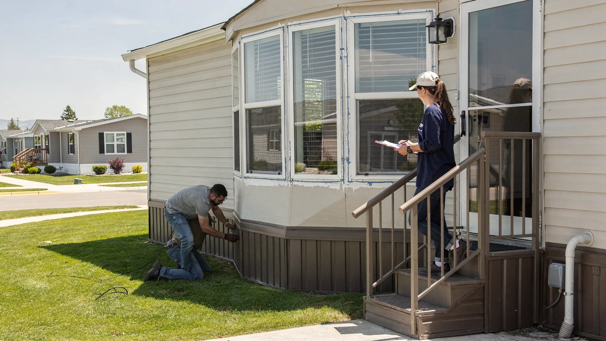 A renter doing an exterior walkaround of a manufactured home, checking skirting panels, looking at window trim and caulking, and inspecting the entry steps and handrail on a sunny day in a manufactured home community.