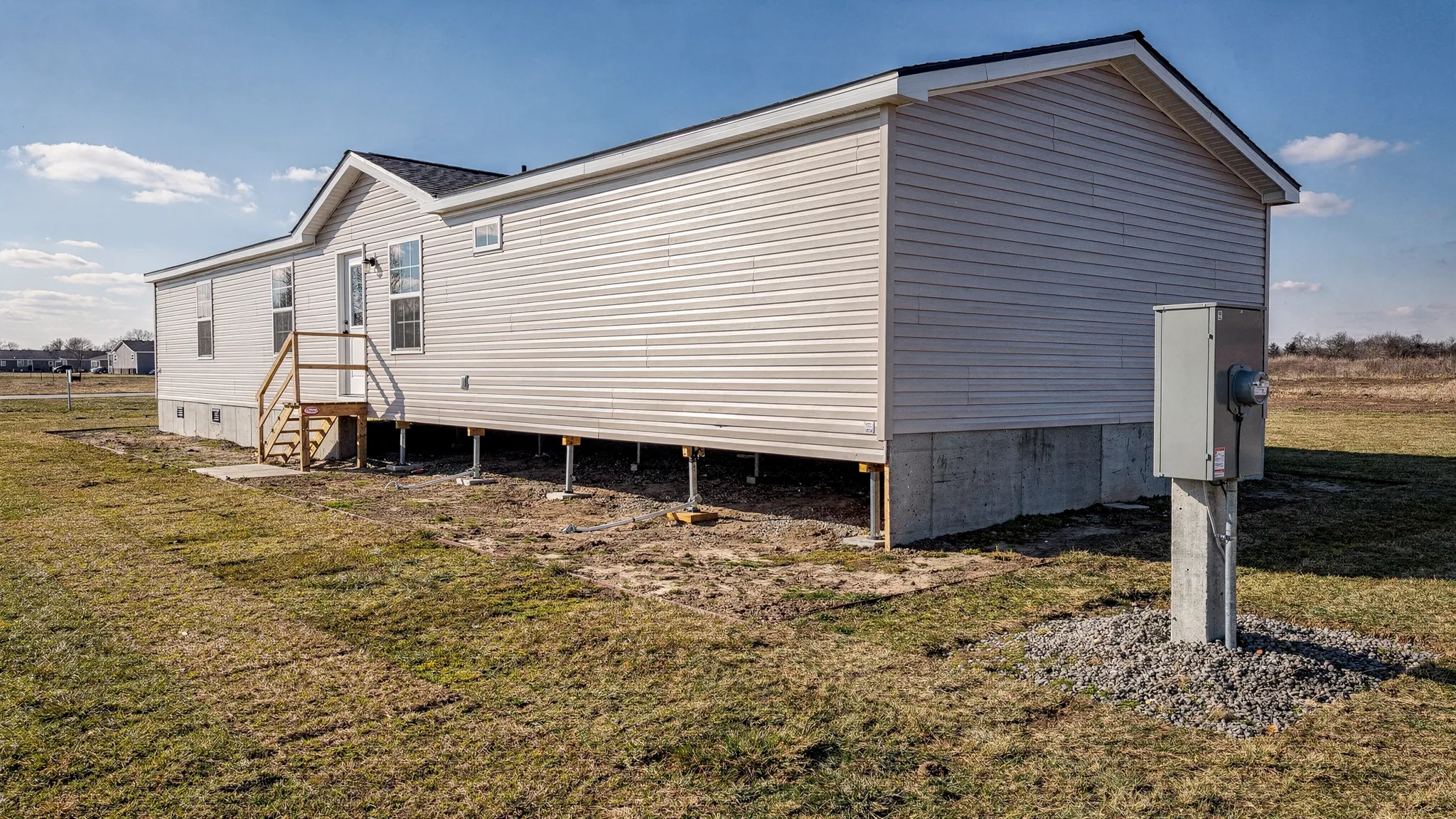 A manufactured home set on a permanent foundation with visible tiedown points and a simple utility pedestal nearby, photographed from a slight angle on a clear day.