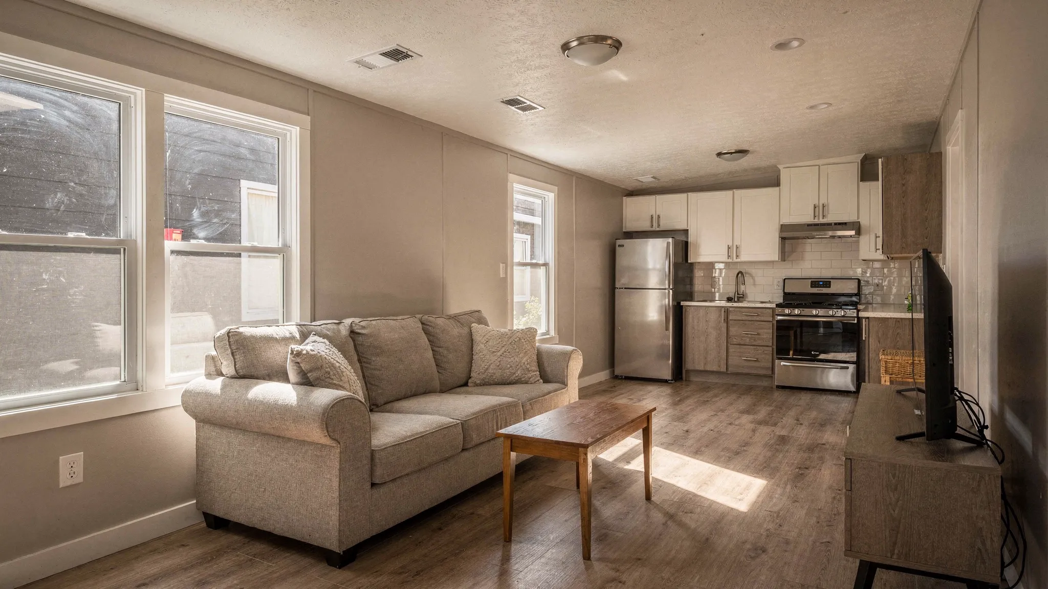 A cozy single wide manufactured home living room with modern finishes, visible insulation-friendly double-pane windows, a compact open kitchen, and ceiling vents indicating central HVAC.