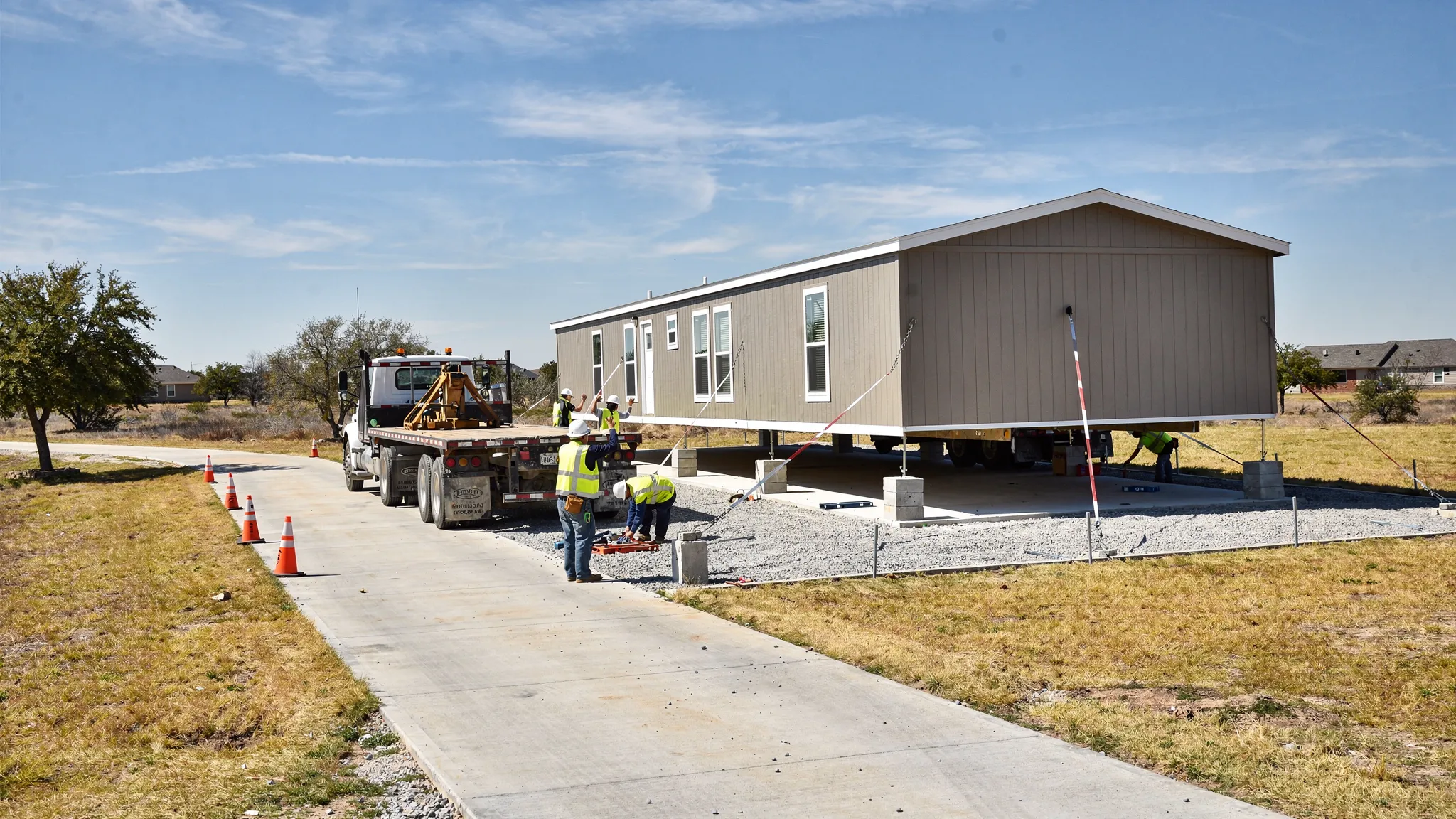 A manufactured home being delivered and set on a prepared pad, with an installer crew guiding placement, tie-down straps visible, and a clear driveway access route on a Texas lot.