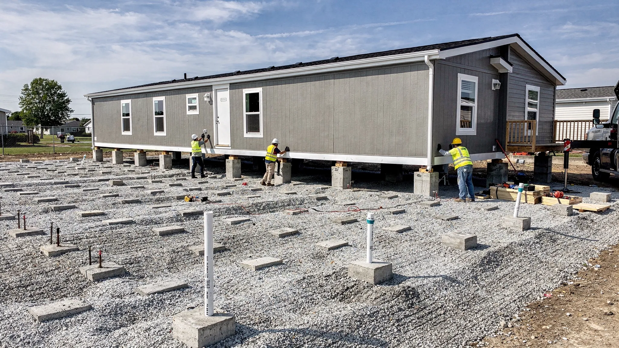 A manufactured home being installed on a prepared pad with visible gravel base, concrete supports, and utility stub-outs nearby, with workers positioning sections for setup.
