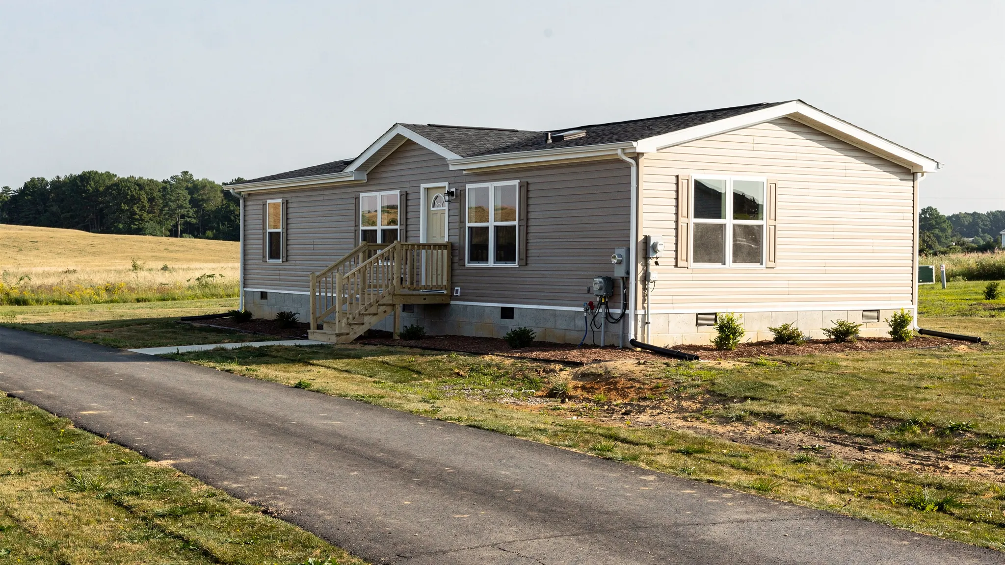 A manufactured home set on a prepared site with a permanent foundation, driveway, utility connections, and a clear view of the surrounding land.