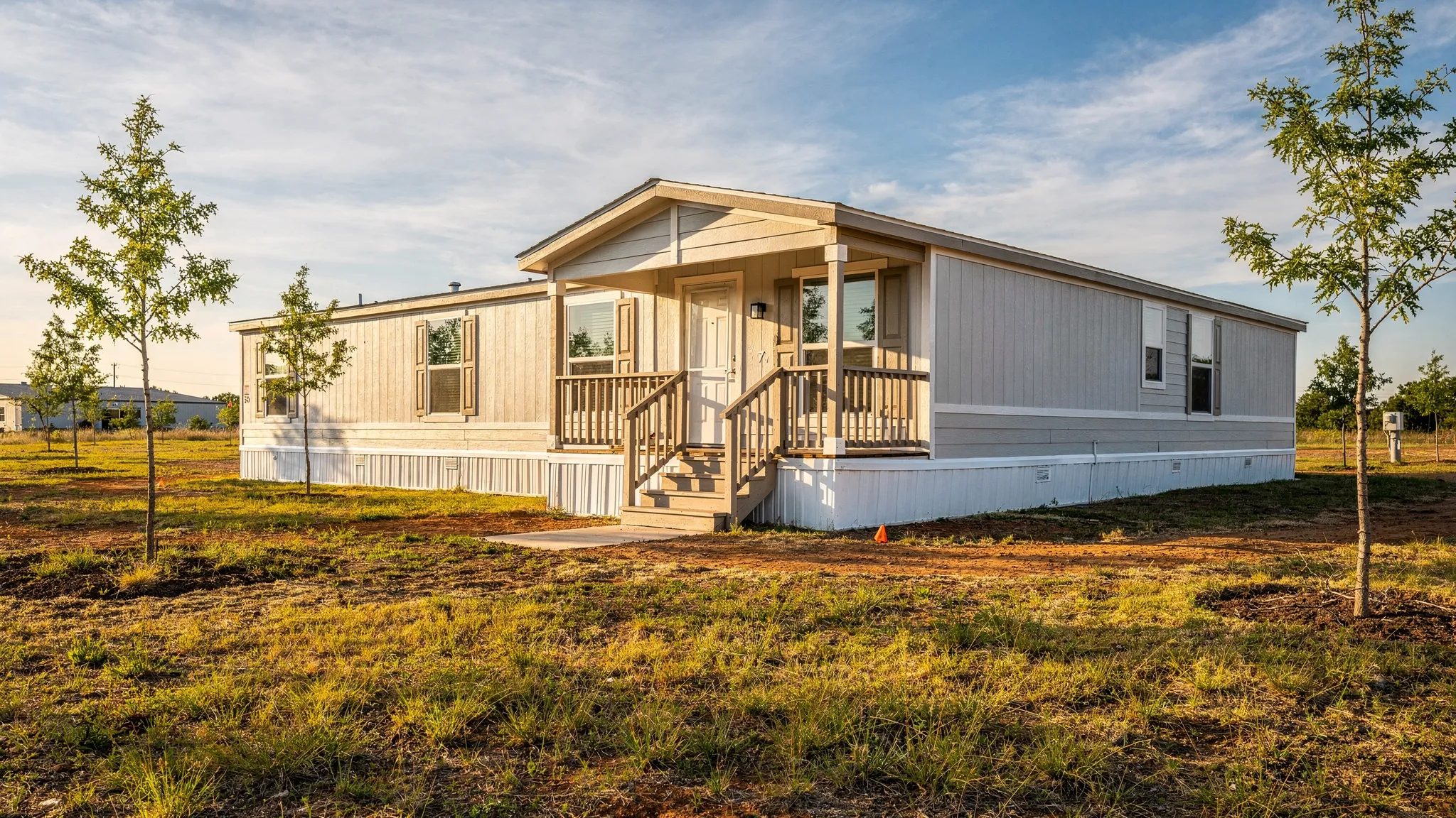 A modern single wide manufactured home with a covered entry, neat skirting, a small front porch, and a sunny San Antonio area lot with native grass and young trees.