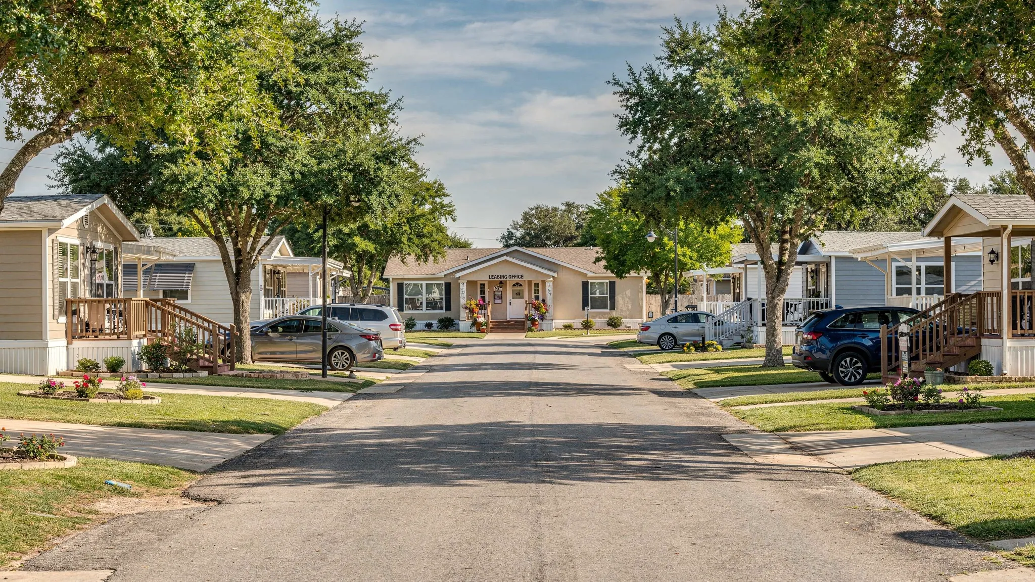 A clean manufactured home community street in the San Antonio area with well-kept homes, driveways, shade trees, and a leasing office in the distance.