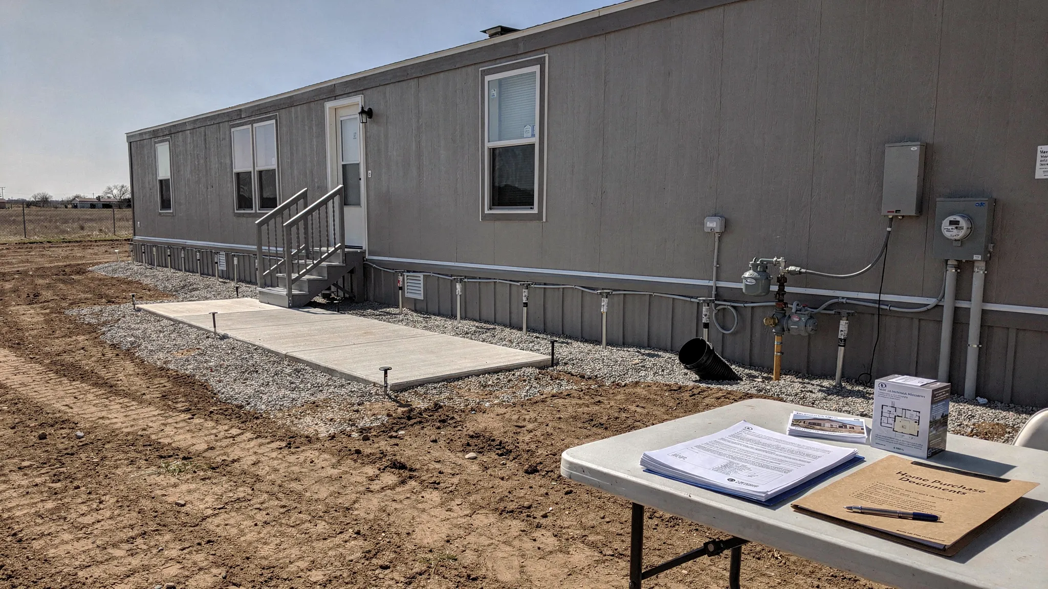 A manufactured home on a prepared Texas lot with concrete pads, utility connections, tie-down anchors, and home purchase documents arranged on a nearby table.