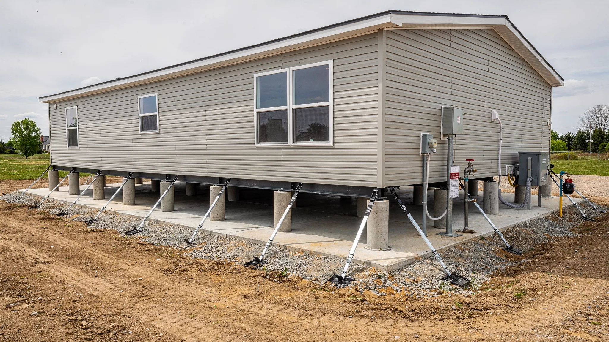 A manufactured home set on a prepared site with visible foundation piers, ground anchors, steel tie down straps, utility connections, and a clear access path around the home for inspection.