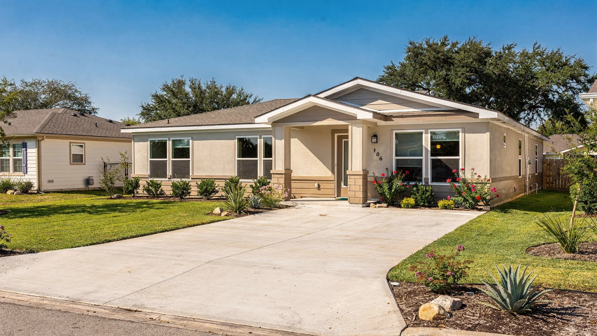 A modern double wide manufactured home in a sunny San Antonio neighborhood with a covered entry, landscaped yard, wide driveway, and warm neutral exterior finishes.