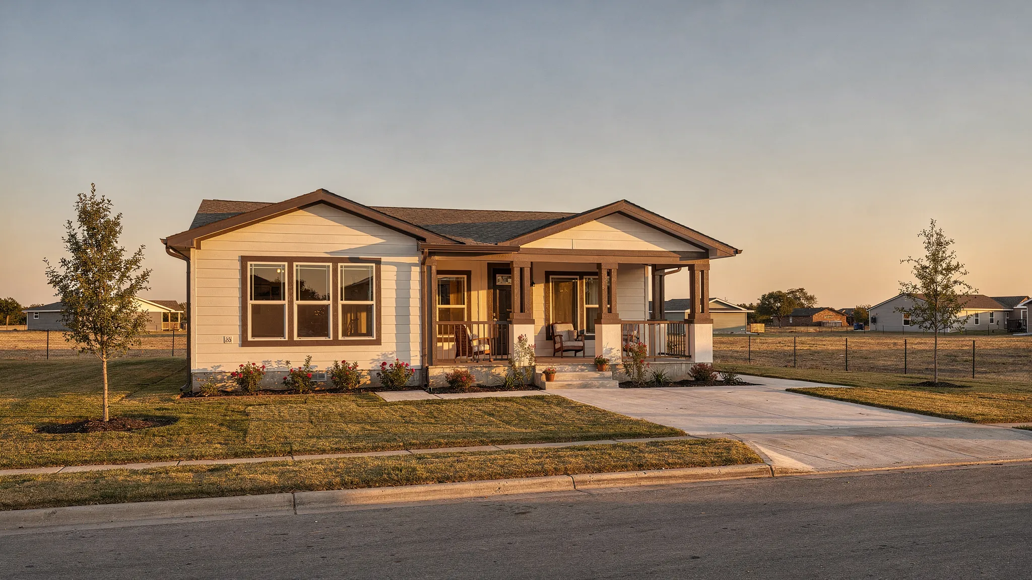 A modern manufactured home in Texas with a covered front porch, simple landscaping, a wide driveway, and warm evening light over an open suburban setting.