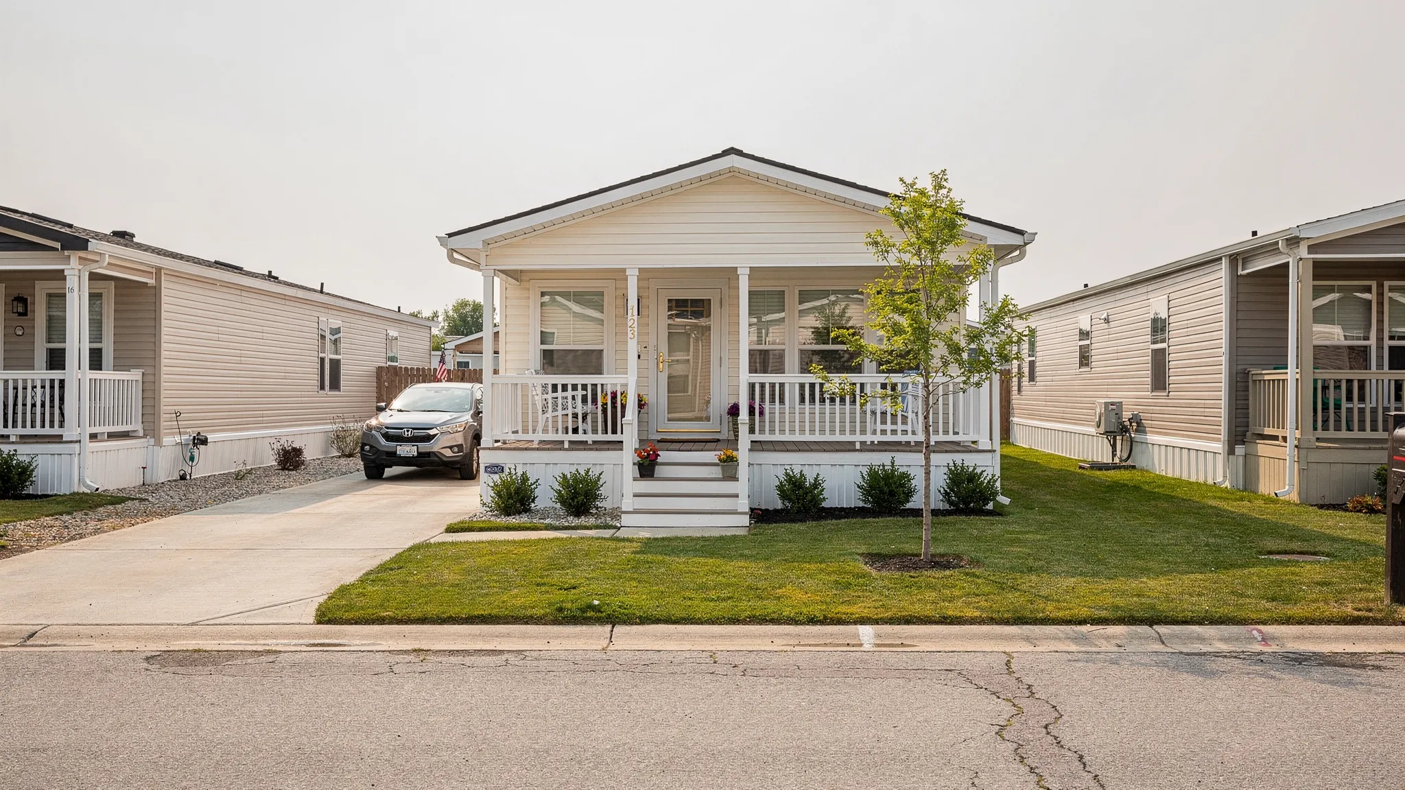 A three bedroom manufactured home with a front porch, driveway, small yard, and neighboring homes in a clean residential community.