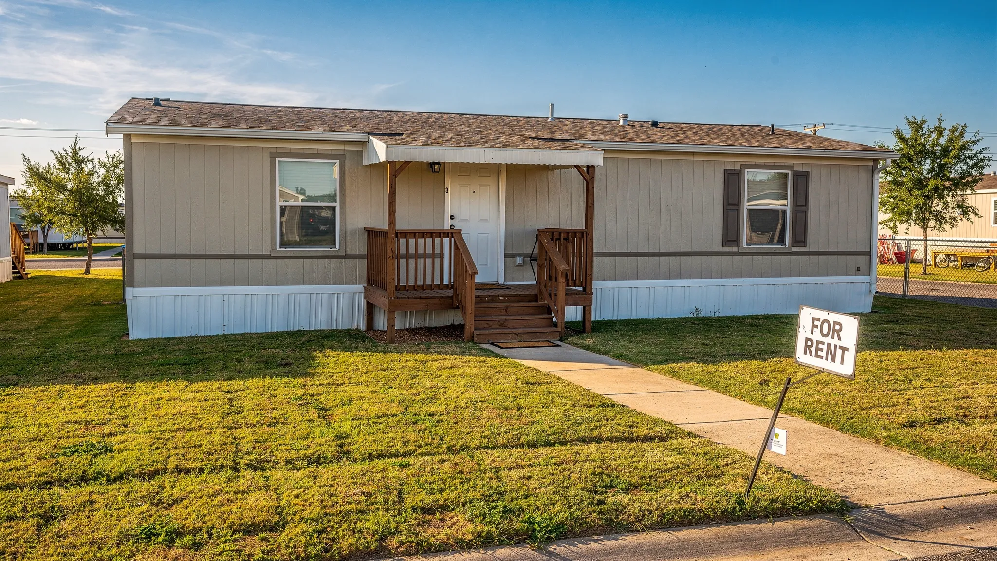 A well-maintained manufactured home with clean skirting, a small front porch, trimmed grass, and a simple “For Rent” sign near the walkway in a sunny Texas neighborhood.