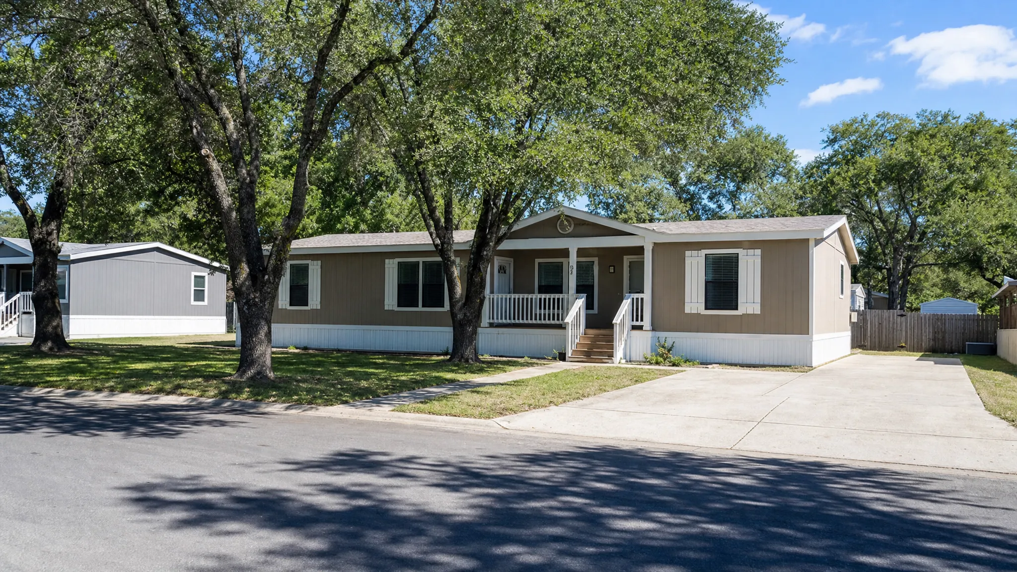 A clean double wide manufactured home in a San Antonio-area community with a small front porch, skirting, driveway, trimmed lawn, and mature shade trees, shown from a wider street view with the home centered and the lot surroundings visible.