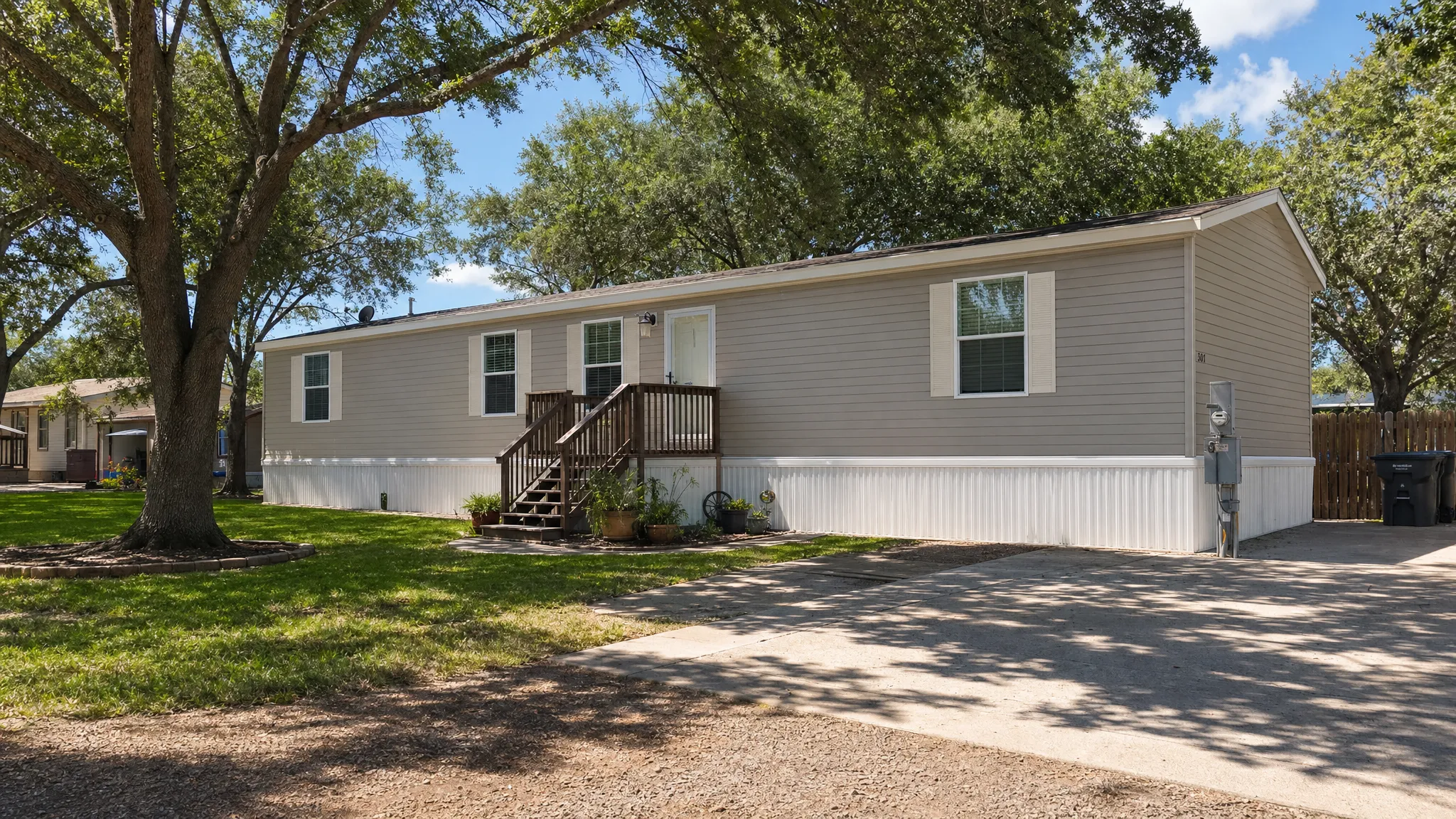 A double-wide manufactured home installed on a prepared residential lot with skirting, front steps, a driveway, utility connections, and a shaded yard in South Texas.