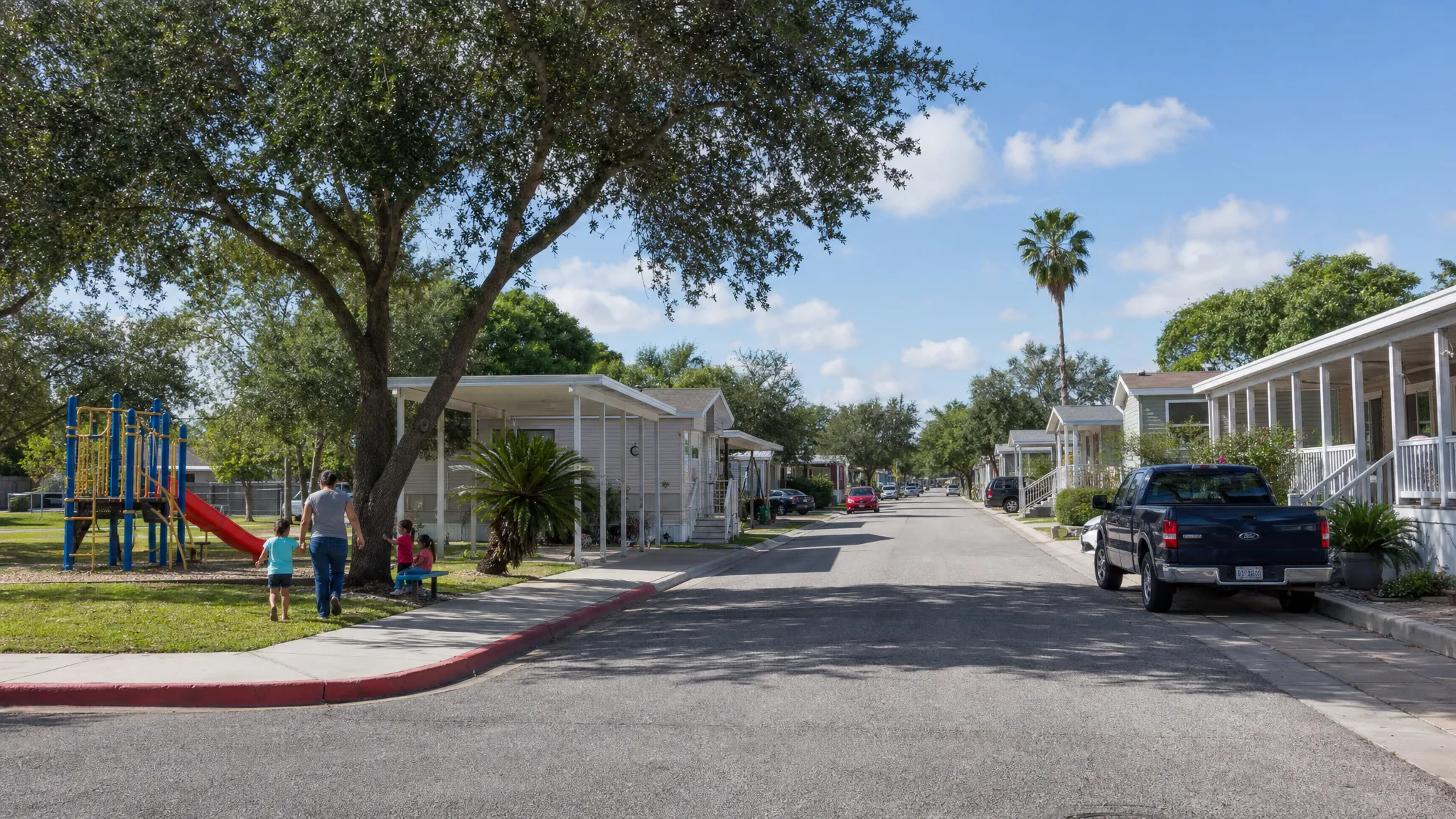 A manufactured home community street in South Texas with well-kept homes, shaded yards, parked vehicles, sidewalks, and residents walking near a small playground.