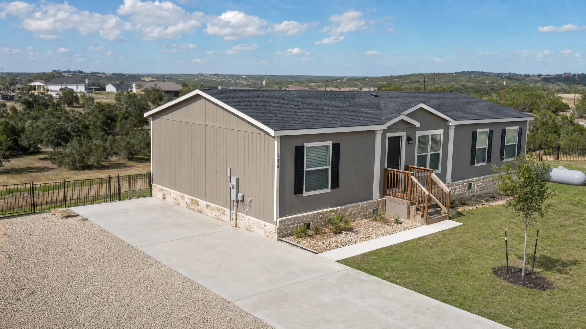 A manufactured home on a prepared residential lot with a permanent foundation, skirting, a driveway, and visible utility hookups, shown from a slightly elevated angle with open Texas landscape in the background.
