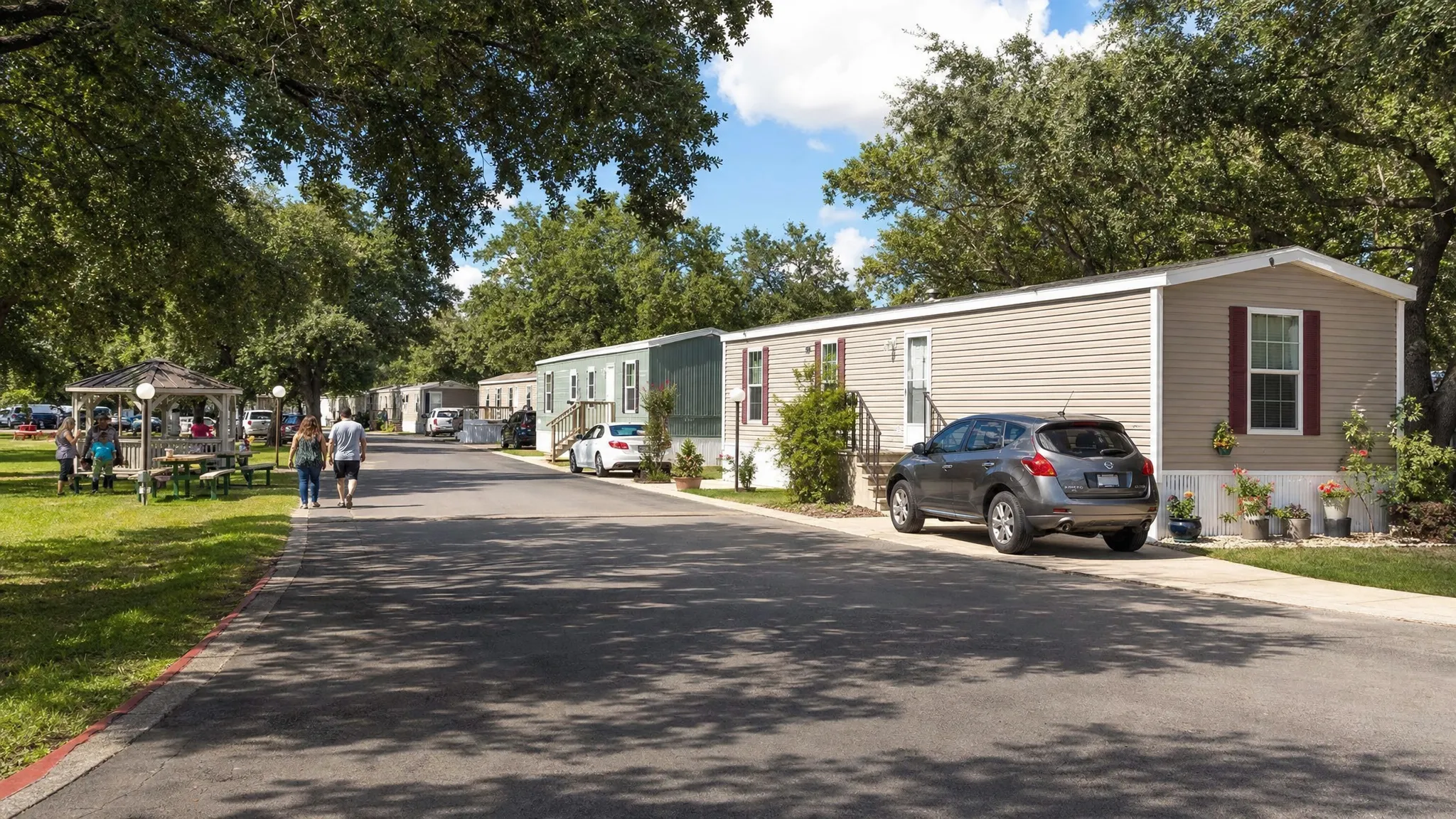 A medium-wide view of a well-maintained all-age manufactured home community in San Antonio, with single-section and double-section homes along a shaded street, parked cars in driveways, and residents walking near a small green common area.