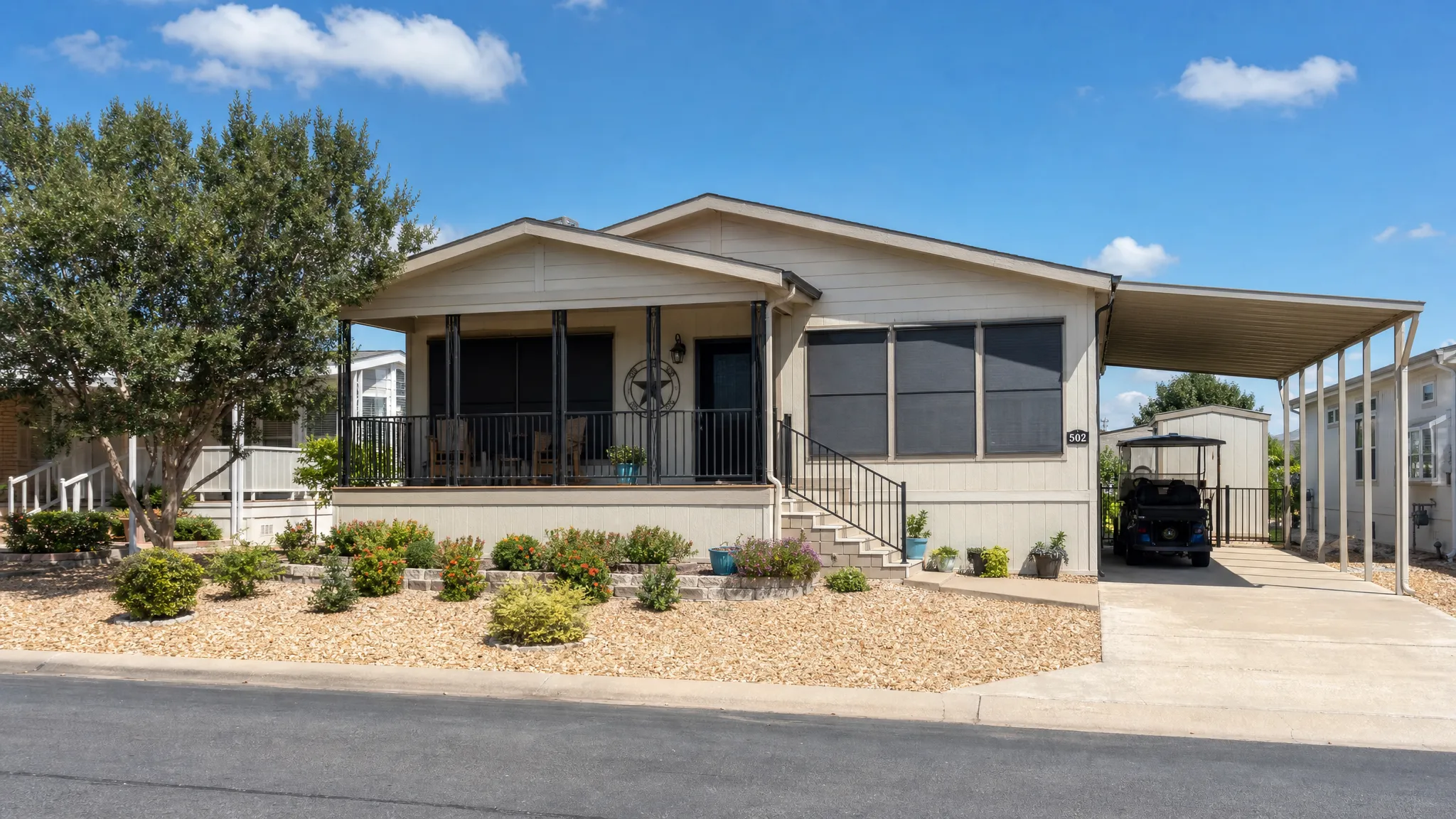 A modern manufactured home in a sunny Texas neighborhood with shaded windows, a covered porch, neat landscaping, and visible features that suggest comfort in hot weather, seen from the street with the full front facade and driveway in view.