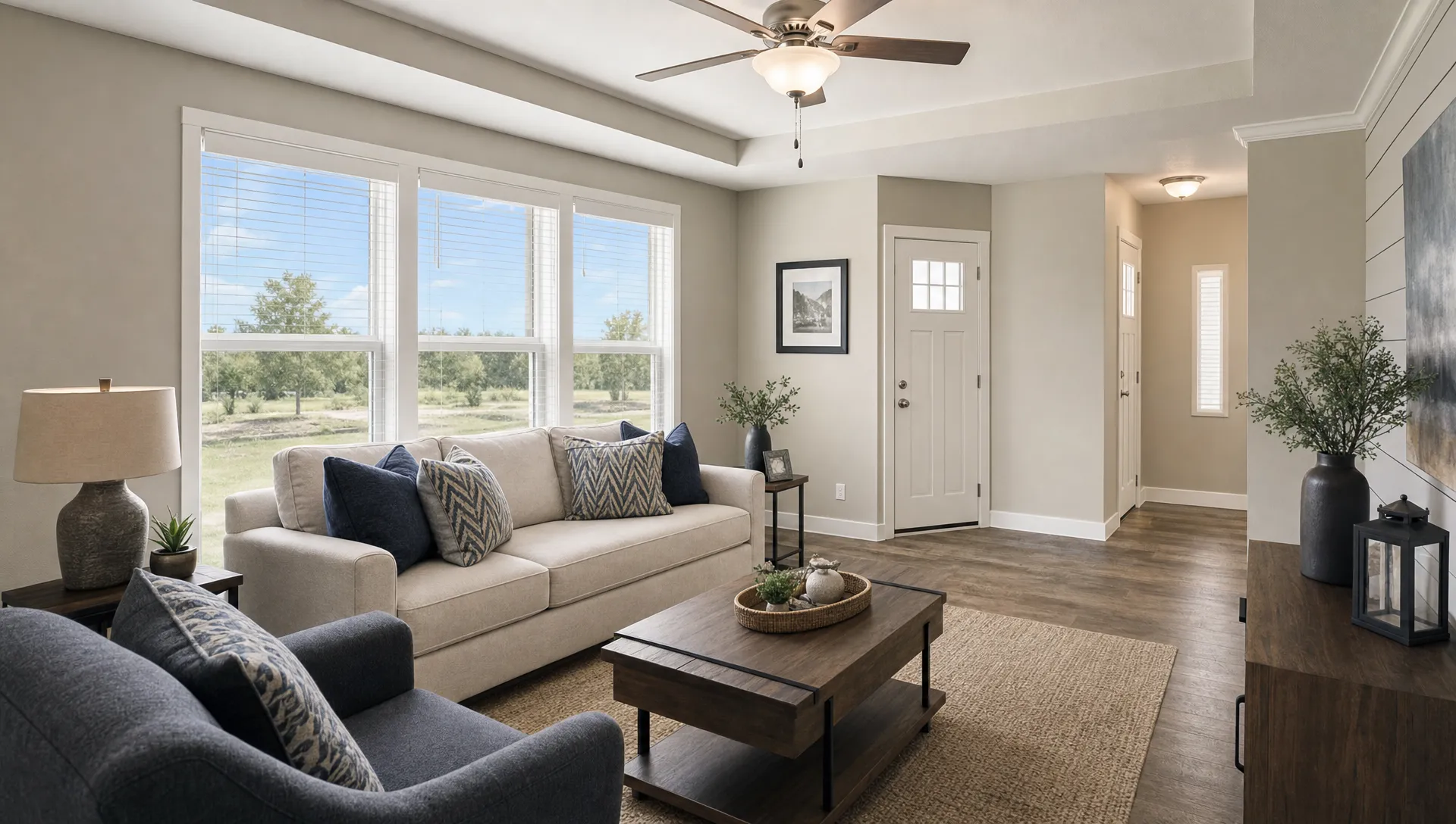 A bright manufactured home living room with large energy-efficient windows, soft natural light, insulated window frames, and a comfortable modern interior designed for hot Texas weather, shown from a corner angle with the windows facing the camera and the seating area arranged below them.