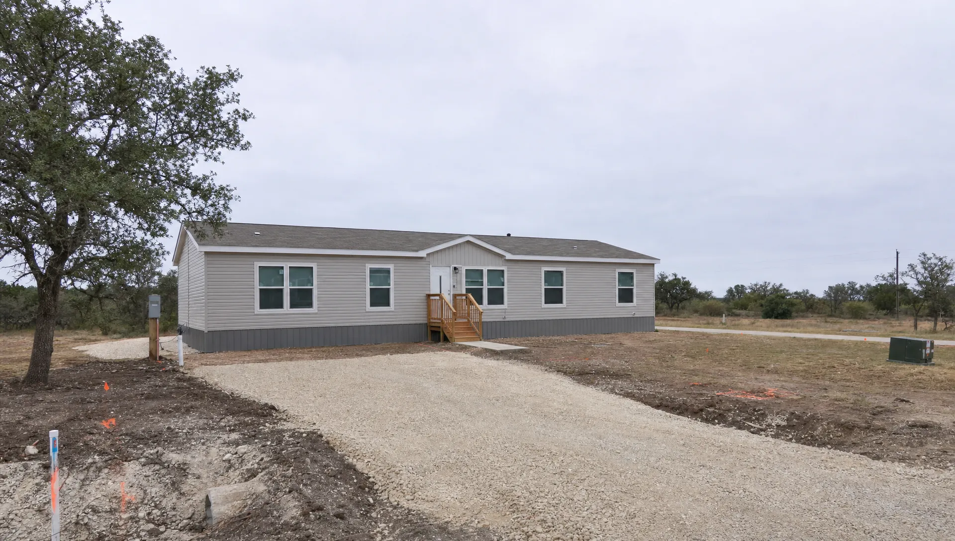 A double wide sits on a residential lot near San Antonio with gravel driveway work, utility markings, and open land around the homesite.