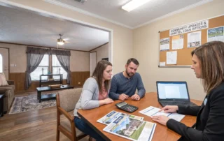 Two adults and a loan officer review manufactured home financing papers beside a laptop in a sales office.