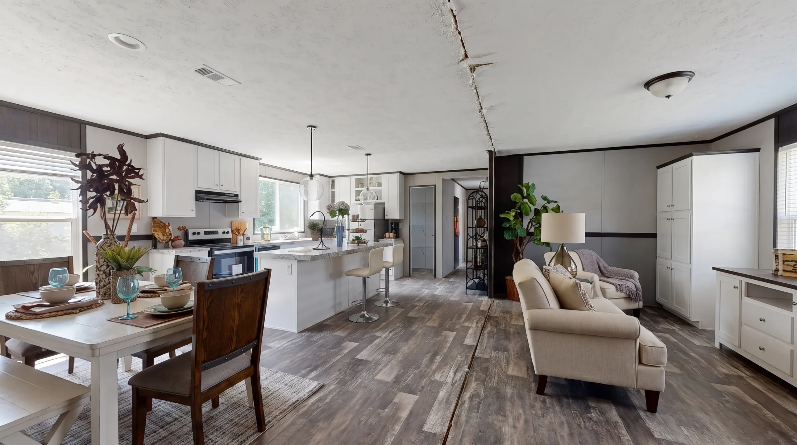 A furnished manufactured home's kitchen, dining area, and living room connect along a clear walkway, with family seating and closed storage nearby.